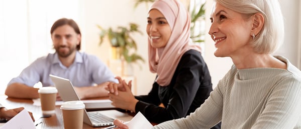 A diverse group of professionals sits around a meeting table in a bright office, smiling and engaged in discussion. A woman wearing a pink hijab speaks while colleagues listen attentively, creating a collaborative atmosphere focused on teamwork, inclusion, and communication.