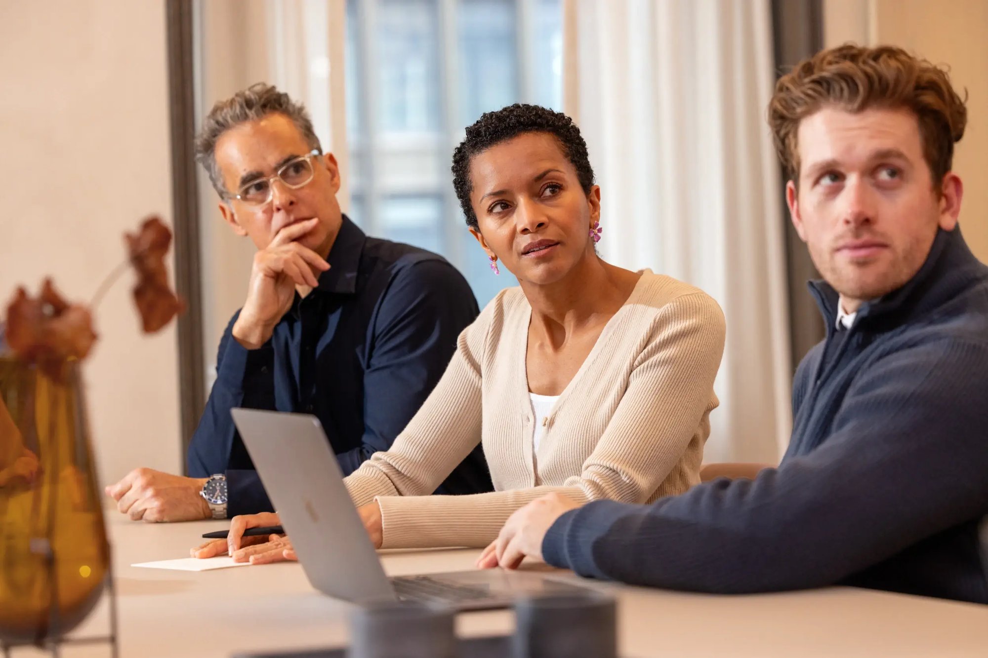 Three colleagues sitting at a table during a meeting, looking attentively toward a speaker off camera while working with a laptop.